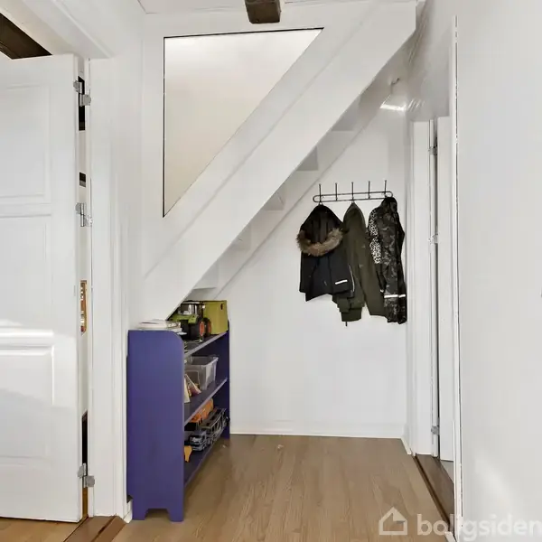 Et hvidt indgangsparti med trægulv og en åben dør. Der er en lilla reol med skuffer og jakker hængende på kroge under en hvid trappe.