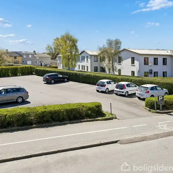 Flere biler holder parkeret på en asfalteret parkeringsplads omgivet af veltrimmede hække. I baggrunden ses boligejendomme under en klar blå himmel med spredte skyer.