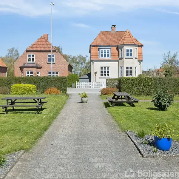 Et hus står for enden af en vej flankeret af græs og haveborde. Røde tage og grønne buske omkranser stedet i et forstadsområde med lys himmel.