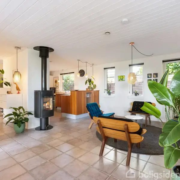 En moderne stue med brændeovn står åben og lys, flankeret af grønne planter. Retro møbler og et rundt sofabord er centralt placeret på gråt tæppe. Lyse vægge og fliser kompletterer rummet.