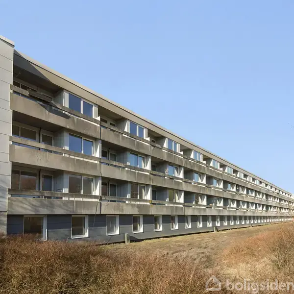 Et langt, gråt lejlighedskompleks står stille mod en klar, blå himmel omgivet af tørre buske. Der er flere etager med balkoner og store vinduer.