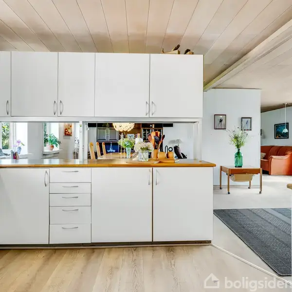 Køkken med hvide skabe og træbordplade. På bordet står blomster og en kaffemaskine. I baggrunden ses spisestue med træmøbler og en stue med sofaer og store vinduer.