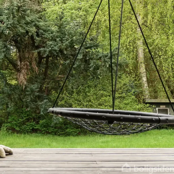 Rund gynge hænger stille, omgivet af en grøn have og træer, med en træterrasse i forgrunden. Huset er sortmalet, og græsset er frodigt grønt.