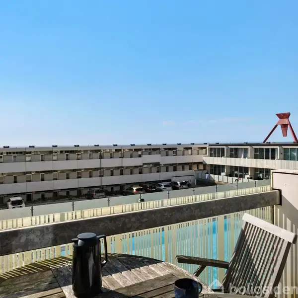 Kaffekande og kop står på et rundt træbord på en altan. I baggrunden ses en række moderne bygninger og en stor rød struktur mod en klar blå himmel.