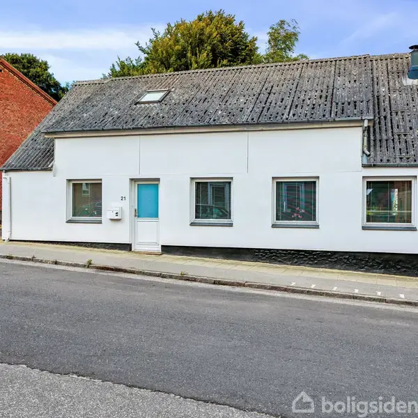 En hvid villa med blå dør og fire vinduer står stille langs en asfalteret vej. Bygningen er omkranset af røde murstensbygninger. Huset har et skråt sort tag.