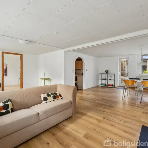 En beige sofa med mønstrede puder står i en lys stue med trægulv. Til højre er der et spisebord med stole og et vindue. En grøn lænestol og et lille bord er i baggrunden.