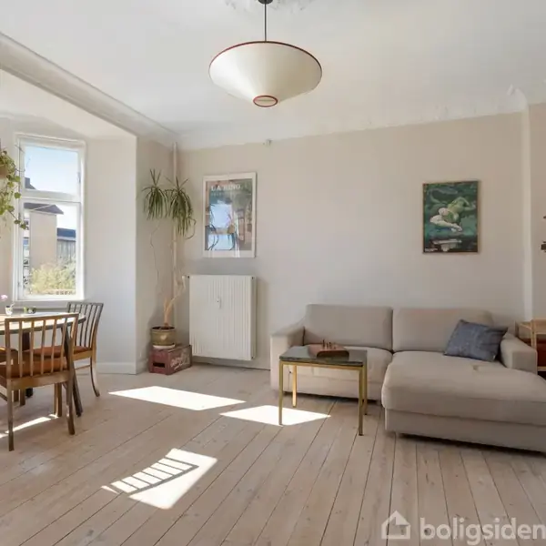 En stue med en lys sofa står i et hjørne, ved siden af et lille bord. Et spisebord med stole ses til venstre nær vinduer. Der hænger planter og billeder på væggene.