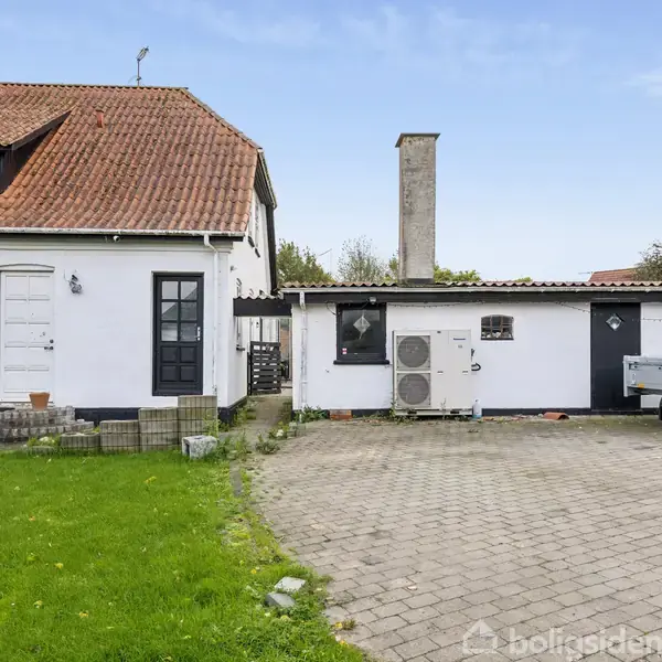 Hus med hvide vægge og rødt tegltag, placeret i en have med grøn græsplæne. Ved siden af huset står en trailer på en brolagt indkørsel.