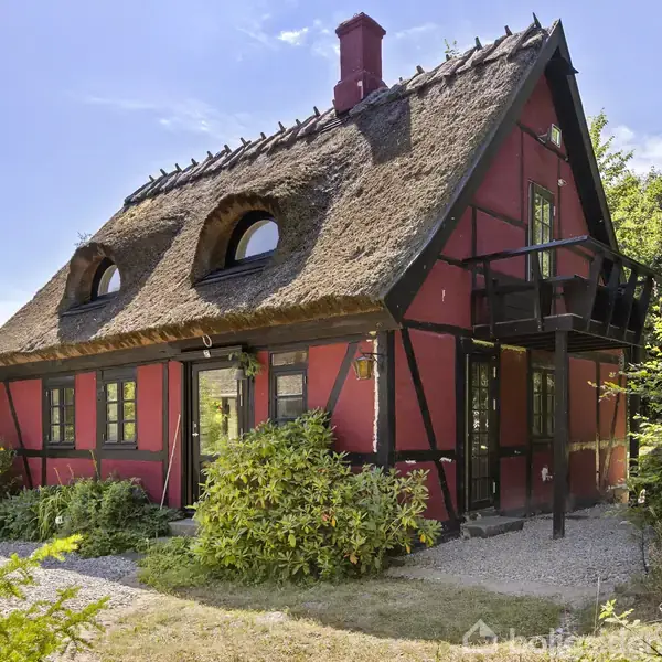 Et bindingsværkshus med stråtag står omgivet af buske og træer. Huset har røde vægge, sorte træbjælker, og en balkon på første sal. Større fliser danner en lille indgangsvej.