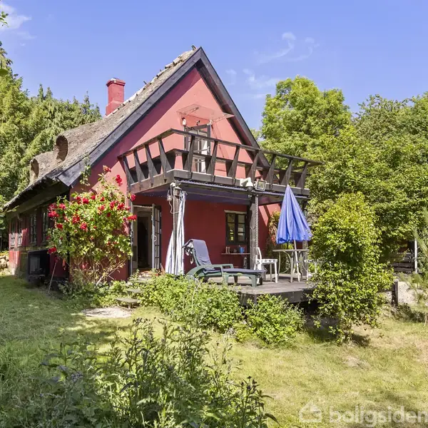Rødt bindingsværkshus med stråtag står stille i en frodig have omgivet af træer, med solrig terrasse og blå parasol.