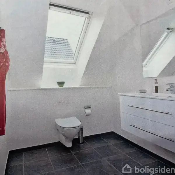 Et badeværelse med hvidt toilet, en vask med skab, og stort spejl. Et ovenlysvindue lader lys ind og afslører et rødt håndklæde hængende. Gulvet er mørkt flisebelagt.