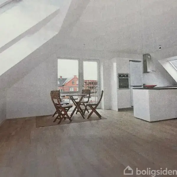 Et spisebord med to stole står i et lyst, skråtloftet køkkenrum med trægulv. Store vinduer og ovenlysvinduer giver udsigt til nogle bygninger.