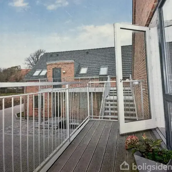 En balkon med træplanker og hvide gelændere er åben mod en trappe indendørs. Omkring er der murstensbygninger med tagvinduer under en blå, let skyet himmel.