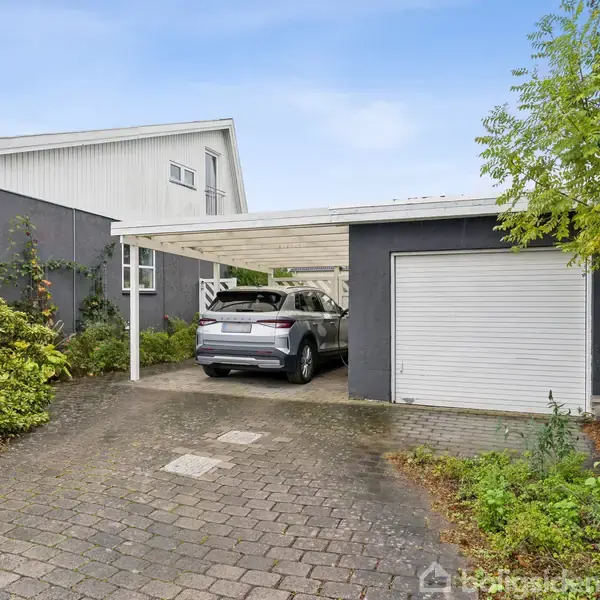 Bilen holder parkeret under en carport ved siden af en sort malet bygning med et hvidt garageport. Indkørslen er brolagt, omgivet af grønne buske og træer.