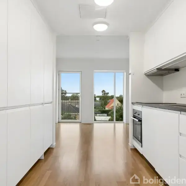 Et moderne køkken med hvide skabe og stålovn strækker sig langs en smal passage. Rummet har trægulv, og store vinduer i enden giver udsigt over et grønt boligområde.