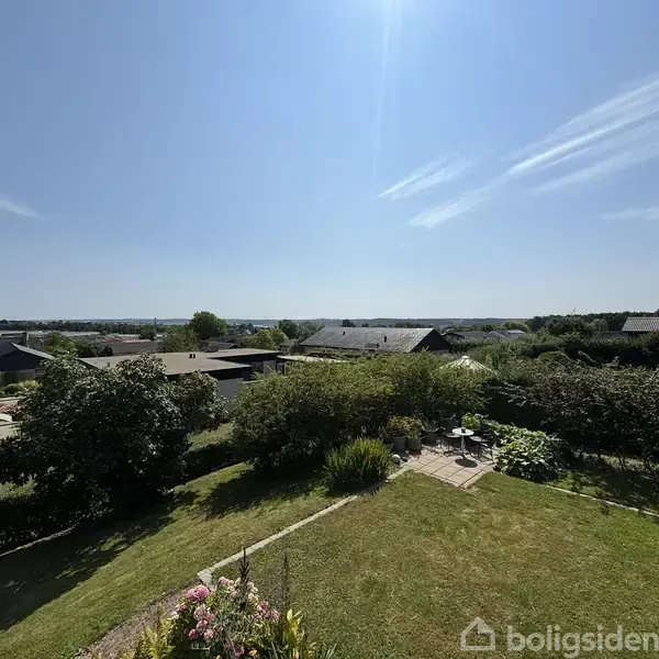 Grøn have med buske og en lille terrasse har udsigt over hustage i et solrigt boligområde. Blå himmel med lette skyer giver en klar og lys atmosfære.