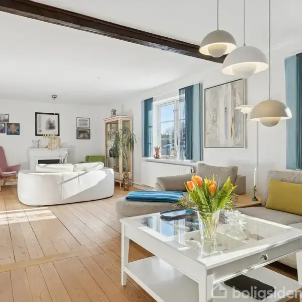 Stue med en hvid sofa og træbord fyldt med tulipaner, omgivet af kunst på væggene. Store vinduer slipper lys ind, og trægulve dominerer i en lys og moderne indretning.