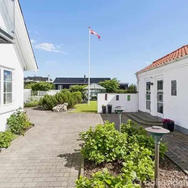 En dansk flagstang står i en solrig gård mellem to hvide bygninger med røde tegltage. Pæne grønne planter omgiver flisebelægningen, og himlen er klar og blå.