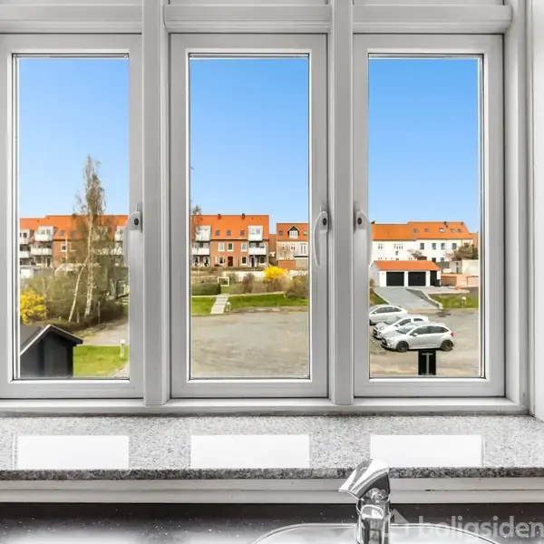 Et køkkenvindue med udsigt over en parkeringsplads og flere bygninger med røde tage i baggrunden. Sollys skinner gennem vinduet, og en enkelt vandhane ses i forgrunden.