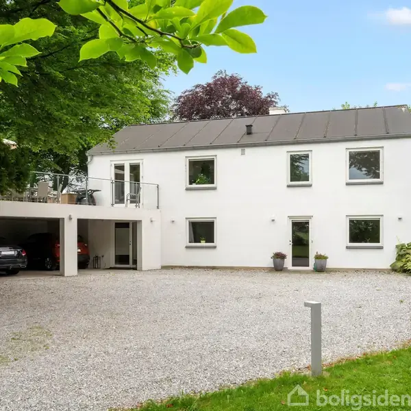 Toetagers hvidt hus med store vinduer, omgivet af træer og buske. En grusindkørsel fører til en carport med biler. Foran huset står blomsterkrukker og en parasol.