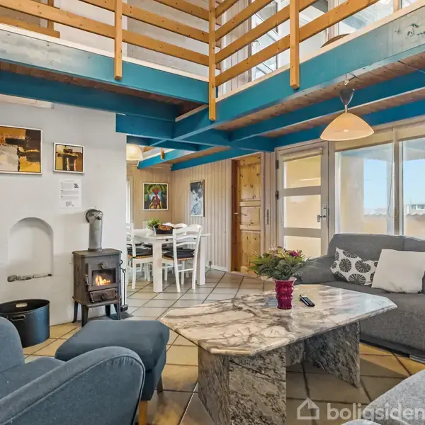 En hyggelig stue med grå sofa og marmor sofabord. En brændeovn opvarmer rummet. Ovenpå ses en træloftskonstruktion. Store vinduer giver udsigt til en solrig terrasse.