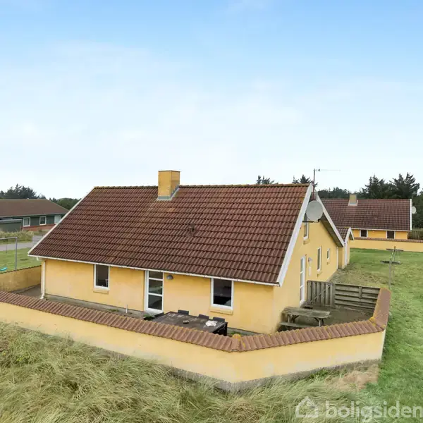 Et gult murstenshus med rødt tegltag står stille ved siden af en lille indhegnet have i et grønt område. Andre huse ses i baggrunden under en blå himmel.