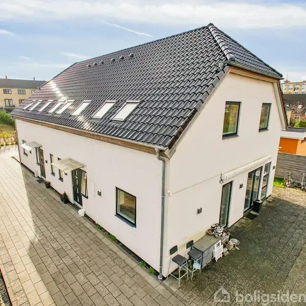 Et moderne, hvidt to-etagers hus med sort tag står på en flisebelagt terrasse, omgivet af grønne områder og andre huse i et boligkvarter.