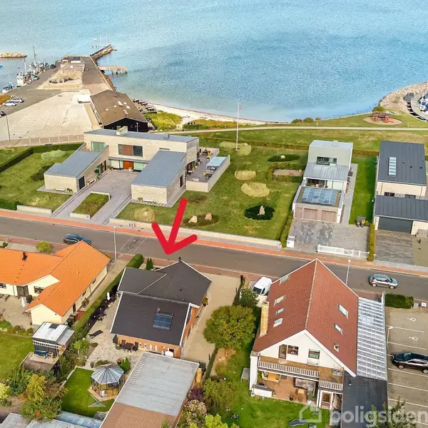 Et hus med sort tag er markeret med en rød pil. Huset ligger i et boligområde tæt på kysten. Omkringliggende huse har forskellige tagfarver. I nærheden er en marina med både.