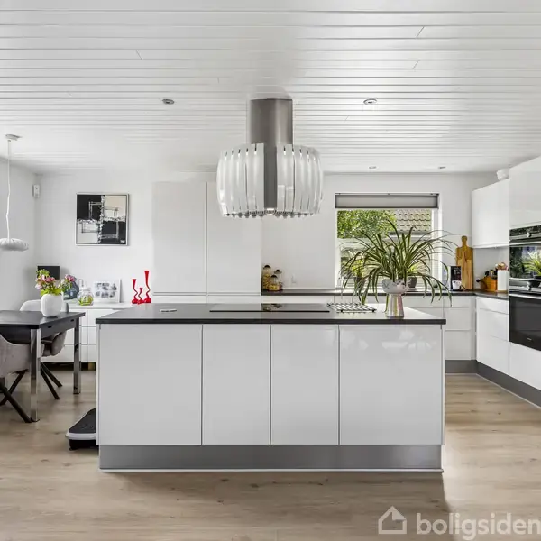 Moderne køkken island med hvidglans skuffefronter står centralt, omkranset af hvide skabe og indbyggede apparater. Et bord med stole og en stor lampe hænger over øen. Store vinduer giver lys.