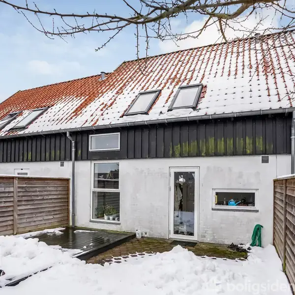 Rækkehus med rødt tegltag og flere ovenlysvinduer ses fra baghaven dækket af sne. Et hegn omringer området, og et lille bassin er i midten.