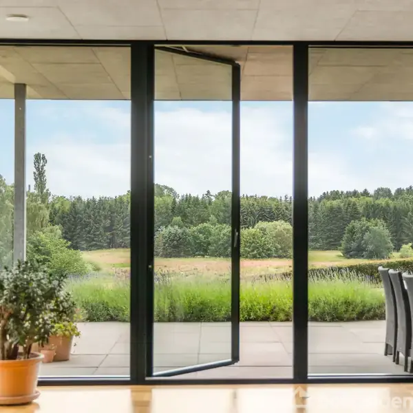 Store glasdøre er åbne mod en terrasse med udsigt til en frodig have og skovbeklædte områder. Indendørs står en grøn plante i en potte ved det åbne rum.