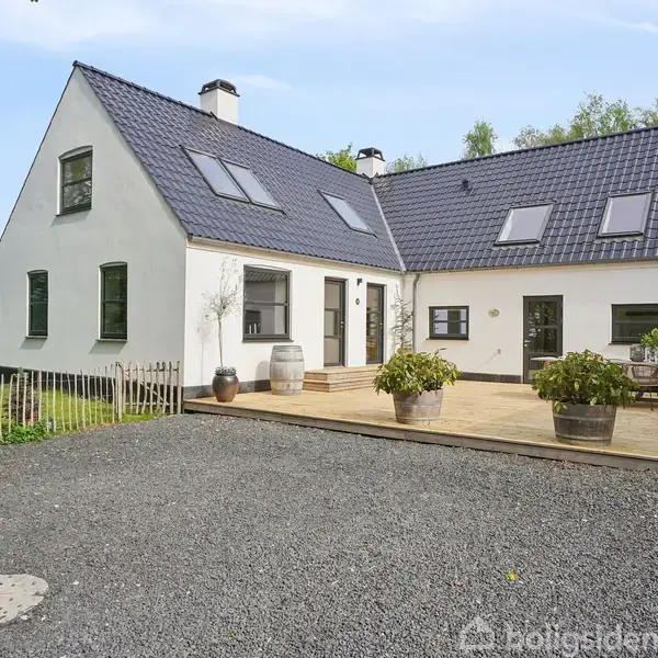 Hvidt hus med blå tagsten, står stille på en grusbelagt gårdsplads, omgivet af frodige træer. Terrasse med blomsterpotter tilføjer en hyggelig atmosfære i det naturlige miljø.