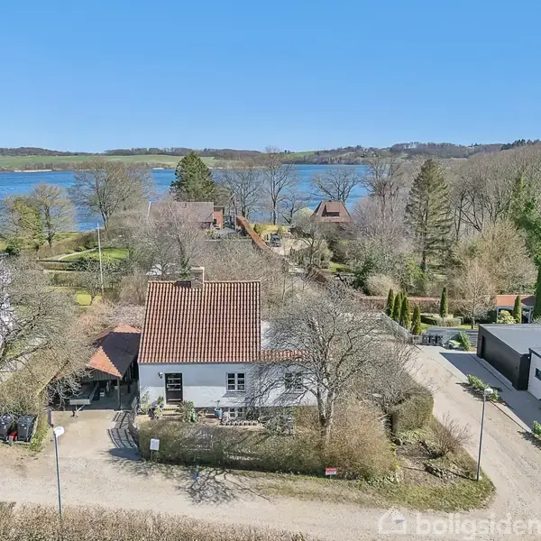 Et hvidt hus med rødt tag står blandt træer og buske ved en grusvej. I baggrunden ses en sø og skovområde under en klar blå himmel.