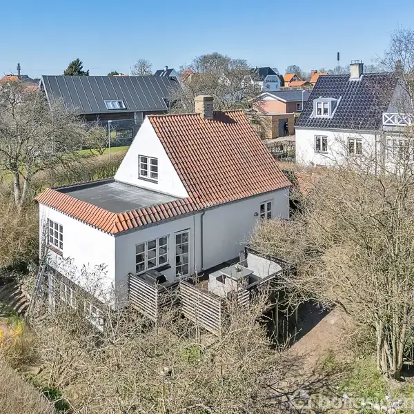Et hvidt hus med rødt tegltag har en træterrasse. Omgivet af træer og buske, står det i et villakvarter med andre huse synlige i baggrunden.