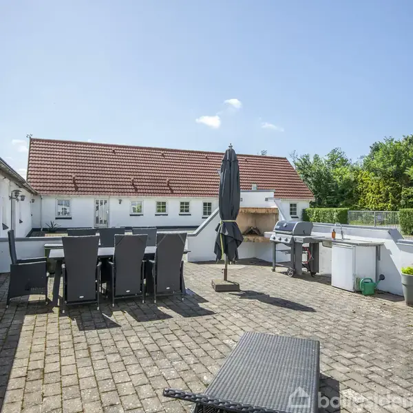 Terrassemøbler står stille på en stor solrig gårdsplads med murstensbelægning. En grill og parasol supplerer scenen. Bygninger med røde tagsten omkranser området, mens grønne hække udgør baggrunden.