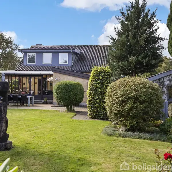 Et hus med to etager står i en velplejet have. En terrasse med møbler er ved huset, omgivet af træer og buske. I forgrunden er en abstrakt træskulptur.