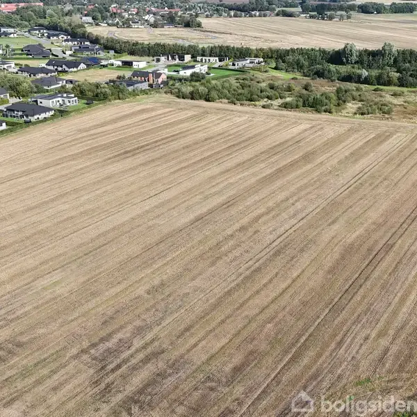En stor, åben mark ligger i forgrunden uden aktivitet. Omgivet af et landligt område med spredte huse og grønne træer.