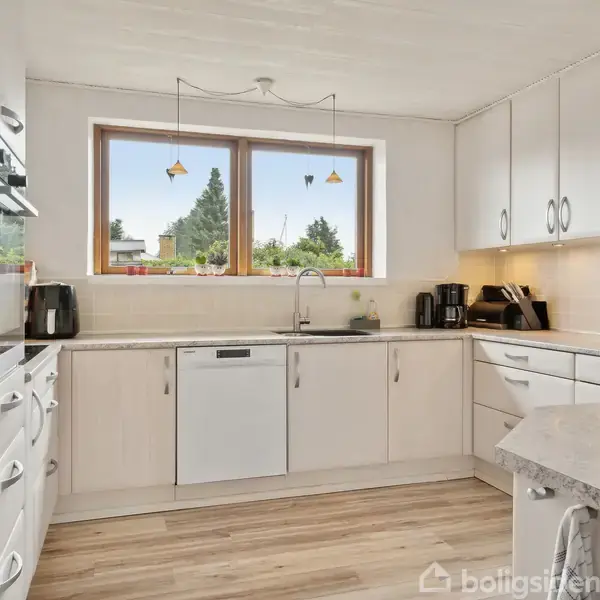 Et moderne køkken med hvide skabe og apparater, herunder ovn og opvaskemaskine. Rummet er lyst med stort vindue, som giver udsigt til grønne træer udenfor.