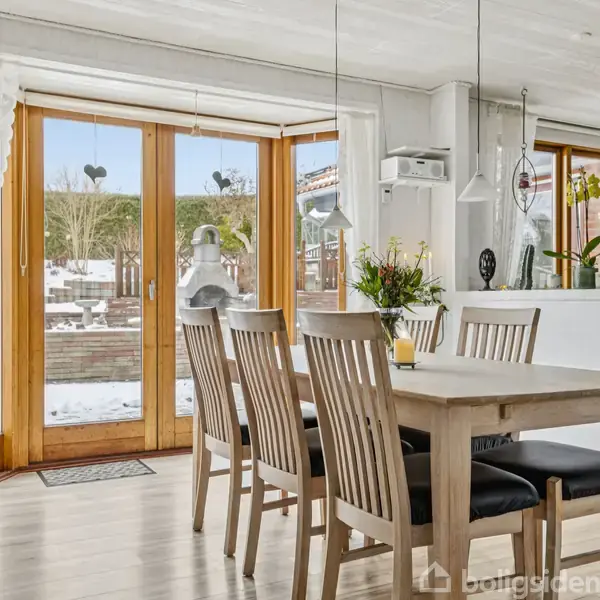 Træbord omgivet af fire stole står i et lyst, skandinavisk spisestueområde. Store vinduer viser en snedækket have. Bordet er dekoreret med en vase med blomster, og moderne lamper hænger fra loftet.