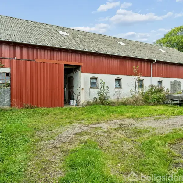 En stor lade med røde og hvide vægge står åben og tom. Den omgivende gårdsplads er fyldt med græs og grus, omgivet af træer og buske.