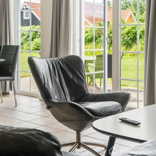 En sort lænestol står ved siden af et sofabord med en fjernbetjening, placeret i en lys stue med store vinduer og udsigt til en have.