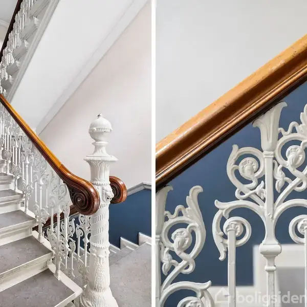 Trappeopgang med detaljeret, hvidt smedejernsgelænder, der snor sig opad. Trin er belagt med gråt tæppe. Blå vægge danner baggrund.