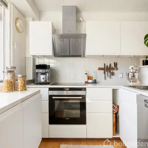 Et minimalistisk køkken med hvide skabe. En komfur med ovn er centralt placeret under en emhætte. Til venstre står glasbeholdere med mad. Et vindue lader lys strømme ind.