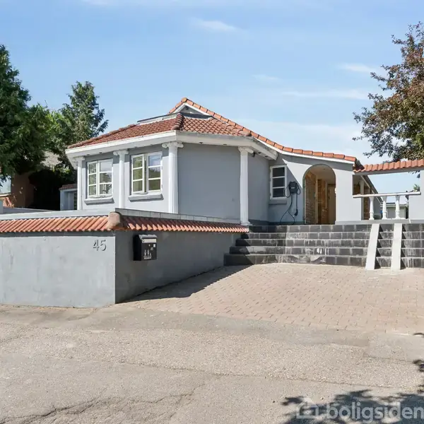 Et gråt hus med rødt tegltag står på en forhøjning, tilgængelig via en trappe. Nummer 45 er malet på murværket. Omgivet af træer og klar himmel.