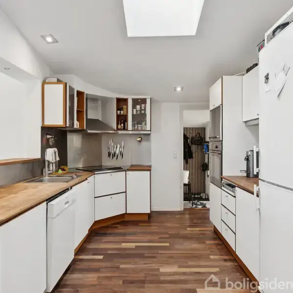 Et lyst køkken med hvide skabe og træbordplader. Køleskab til højre, overskabe til venstre. Rummets trægulv strækker sig ind mod en åben gang.