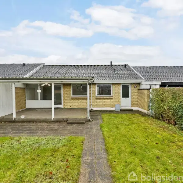 Rækkehus med terrasse i baghaven omgivet af hække. Huset har gule mursten og et gråt tag. Stien leder fra græsplænen til terrassen, der har en overdækning.