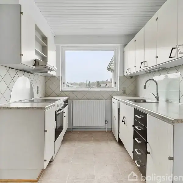 Et lyst køkken med hvide skabslåger og grå bordplader står tomt. Køkkenet har en vask og en komfur, med flisebelagt backsplash. Et stort vindue giver udsigt til omgivelserne.