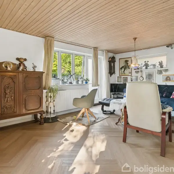 En stue indeholder en vintage kommode, stole og sofabord med bøger. Store vinduer slipper lys ind, og planter samt kunstværker dekorerer rummet. Hyggelig, traditionel indretning skaber en afslappet atmosfære.