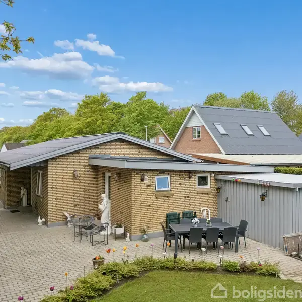 En gul murstensbygning med terrasse er omkranset af en have. Terrassen har et sort bord med otte stole, og skur i baggrunden. Himmel med få skyer og grønne træer.