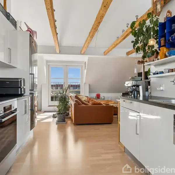 Et moderne køkken-alrum med hvide skabe strækker sig mod en lys stue med sofa og planter. Træbjælker i loftet tilføjer varme. Vinduer slipper sollys ind.